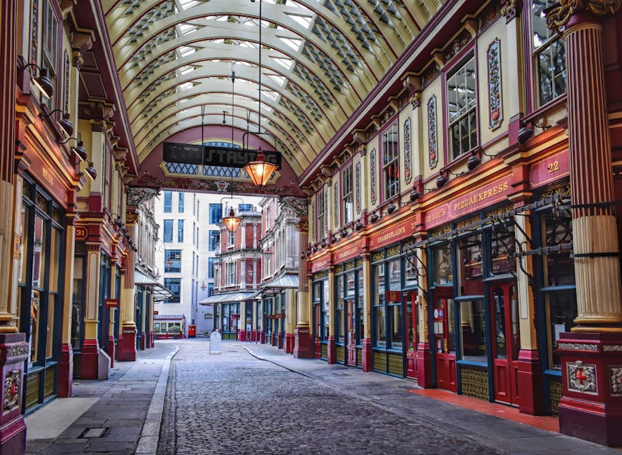 The image depicts an indoor shopping arcade with ornate architectural details, including a high, vaulted ceiling with glass panels allowing natural light to illuminate the space. The corridor is lined with small retail units on both sides, featuring decorative mouldings, painted in shades of red, yellow, and gold, with shop signs displaying various names such as 'Pizza Express' and 'Chocolatiers.' The shops have large glass windows and doors, some with metal grilles, and the pavement is paved with cobblestones. At the far end, the street opens to an outdoor area with taller modern buildings visible in the background. The environment appears clean and well-maintained, with subtle indications of relocation activity in the context of a house removal, as Man with Van Mayfair might organise furniture transport and packing logistics, although no moving equipment or boxes are directly visible in this particular shot.