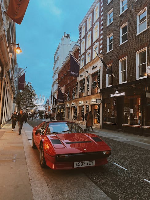 A red sports car with a sleek, low-profile design is parked on a narrow street in Mayfair, London, near the entrance of a building with large windows and black signage. The vehicle's smooth, shiny surface and distinctive shape suggest it is a classic or vintage model, positioned close to the pavement on the right side of the image. Behind the car, there are pedestrians walking along the sidewalk, some carrying shopping bags or engaged in conversation. The street is lined with multi-story buildings featuring traditional brick facades, large windows, and decorative flags hanging from the facades, indicating a commercial or high-end shopping area. Overhead, the sky is transitioning from daylight to evening, with a bluish hue, and street lighting begins to illuminate the scene. Visible in the background, a string of decorative lights stretches across the street, enhancing the lively atmosphere typical of Mayfair. This scene might relate to home relocation or furniture transport logistics, which Man with Van Mayfair could facilitate through professional moving services involving careful packing and transportation of valuable items during a move or house removal.