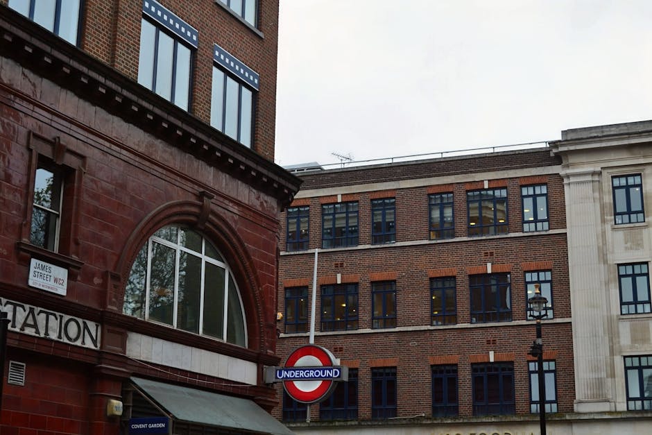 The image depicts an indoor shopping arcade with ornate architectural details, including a high, vaulted ceiling with glass panels allowing natural light to illuminate the space. The corridor is lined with small retail units on both sides, featuring decorative mouldings, painted in shades of red, yellow, and gold, with shop signs displaying various names such as 'Pizza Express' and 'Chocolatiers.' The shops have large glass windows and doors, some with metal grilles, and the pavement is paved with cobblestones. At the far end, the street opens to an outdoor area with taller modern buildings visible in the background. The environment appears clean and well-maintained, with subtle indications of relocation activity in the context of a house removal, as Man with Van Mayfair might organise furniture transport and packing logistics, although no moving equipment or boxes are directly visible in this particular shot.