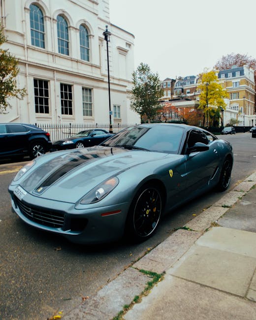 A sleek, dark grey Ferrari sports car with a low profile and aerodynamic design is parked on the street adjacent to a sidewalk. The vehicle features a prominent front grille, angular headlights, black alloy wheels, and the Ferrari emblem on the side panel near the front. The car's tinted windows and smooth, curved surface are visible, with reflections of the cloudy sky and surrounding buildings on its polished exterior. Behind the Ferrari, there are residential buildings with large windows, some with white facades and ornate architectural details, and a row of parked cars along the street. In the background, trees with yellow and green foliage suggest an autumn setting. The scene appears to be in an urban area suitable for luxury vehicle transport during home relocation or moving activities, aligning with services provided by Man with Van Mayfair.