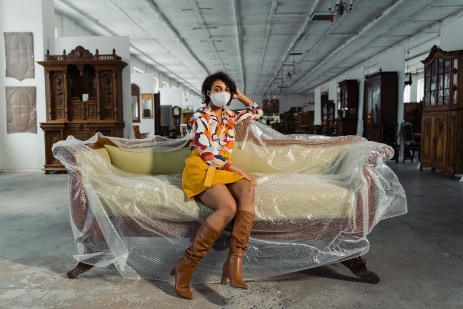 A woman wearing a face mask and a colorful patterned blouse with a yellow skirt and tall brown leather boots is sitting on a vintage sofa that is wrapped in clear protective bubble wrap. The sofa features wooden legs and light-colored upholstery, positioned inside a spacious warehouse with a high ceiling, exposed concrete walls, and large windows allowing natural light. Behind her, several other antique wooden furniture pieces, including cabinets and sideboards, are arranged along the walls, indicating an environment where furniture is prepared for packing, transport, or home relocation. The floor appears to be concrete, and some furniture items in the background are enclosed in wrapping or protective materials, highlighting careful handling and packing processes essential during furniture transport and removals. Man with Van Mayfair, a company specialising in removals, may be involved in this home relocation project which includes packing, loading, and moving antique furniture with care and attention to detail.