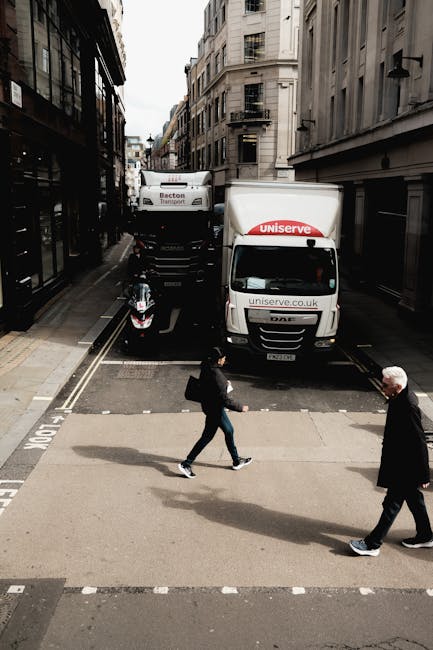 A street scene in an urban area with narrow roads flanked by tall, multi-storey buildings featuring traditional and modern architectural elements. Two white moving trucks, branded with 'Uniserve' and 'Becton Transport,' are parked on the road, with the rear of the trucks extending into the roadway, indicating active loading or unloading operations typical of home relocation or furniture transport. A black motorcycle is parked behind the trucks on the side of the street. In the foreground, two pedestrians—a woman dressed in dark clothing with a backpack and a man in a black coat—are crossing the street on a zebra crossing, walking from right to left. The scene is well-lit with natural daylight, demonstrating the logistical aspects of packing and moving, including vehicle positioning and pedestrian activity near a busy residential or commercial area, with clear focus on the moving process as coordinated by [COMPANY_NAME], supporting services like same-day removals and last-minute move solutions in Mayfair.
