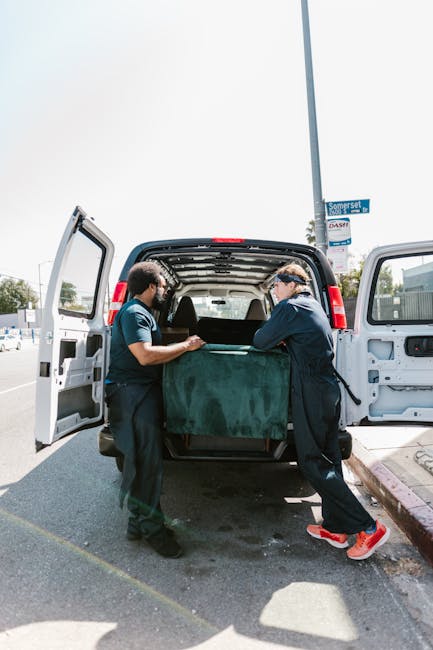Two men dressed in dark clothing and protective glasses are involved in a loading process of a black van parked on a residential street. One man is on the left, holding the edge of a large, green, soft-fabric wrapped item, while the other man, on the right, appears to be guiding or securing the item into the van’s cargo area. The open rear doors reveal additional packed items, possibly furniture or boxes, inside the van, which is situated on a paved road adjacent to a curb and sidewalk. In the background, a street sign is visible alongside a clear sky, indicating a bright day. This scene illustrates a home relocation or furniture transport operation, with the company Man with Van Mayfair providing last-minute removals and moving services in a professional, organized manner, involving careful handling of packing materials and furniture during a same-day move in Mayfair.