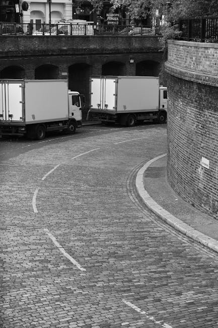 A black and white photograph showing two medium-sized moving trucks parked on a curved cobblestone driveway, with the rear of the trucks facing the camera. The trucks are positioned near a brick retaining wall on the right side of the image, with a metal railing and trees visible above the wall in the background. The driveway has white dashed lines indicating designated parking or loading zones. The scene appears to be part of a home relocation or furniture transport process, with the trucks ready for loading or unloading, and the setting suggests a residential or urban area where professional removals services by Man with Van Mayfair may be utilized to facilitate efficient moving logistics.